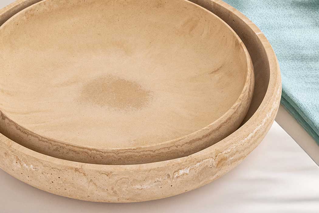 Close-up of two beige, textured travertine stone bowls nested together on a smooth surface, with a soft green cloth nearby, creating a calm, earthy tone.
